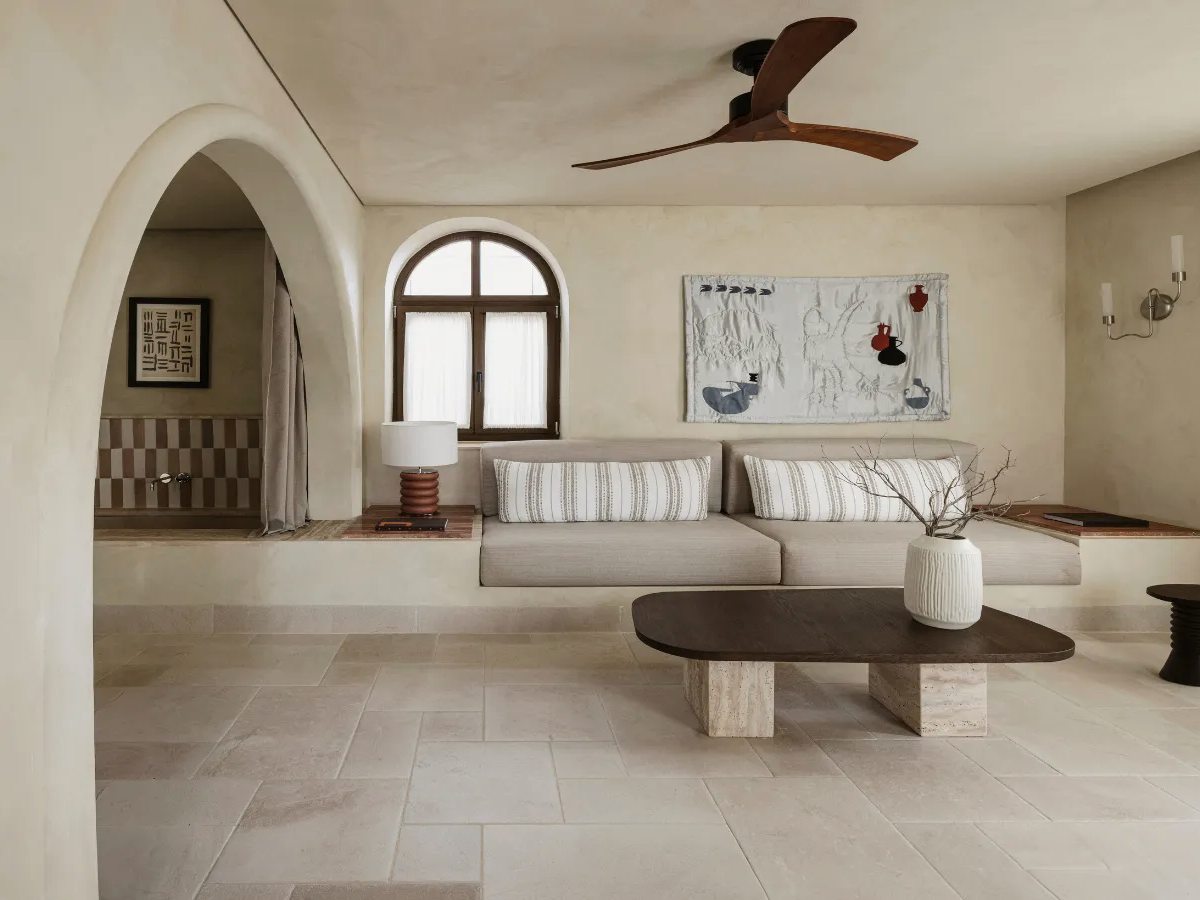 An open-plan suite living area with a low built-in sofa, a stone-topped coffee table, an arched doorway through to a bathroom and a pale stone floor.