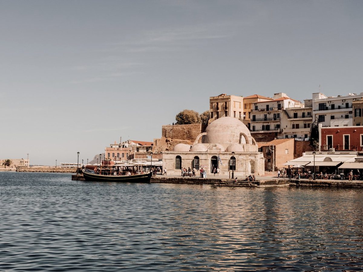 The stone Ottoman mosque and waterfront of Chania's old Venetian harbour under a clear sky, fishing boats moored alongside.