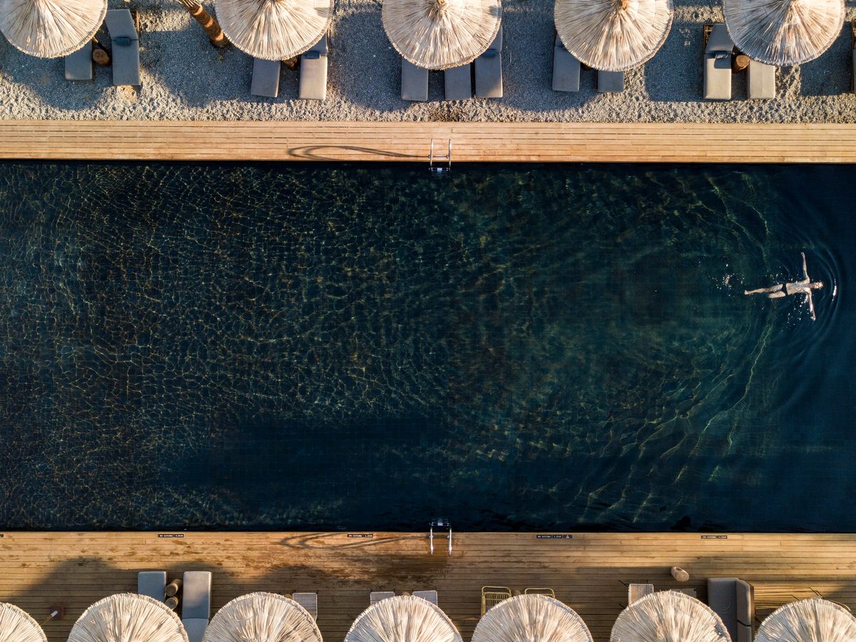 An overhead view of the long black-tiled main pool flanked by wooden decking, thatched umbrellas and paired loungers, with a single swimmer.