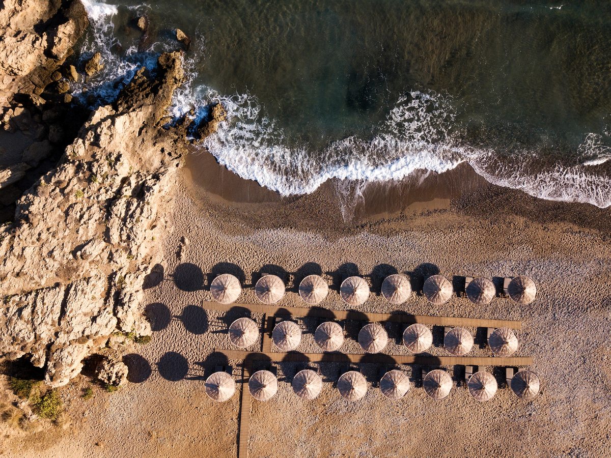 An aerial view of rows of thatched umbrellas and loungers on a grey-pebble beach beside a rocky headland, waves rolling in.