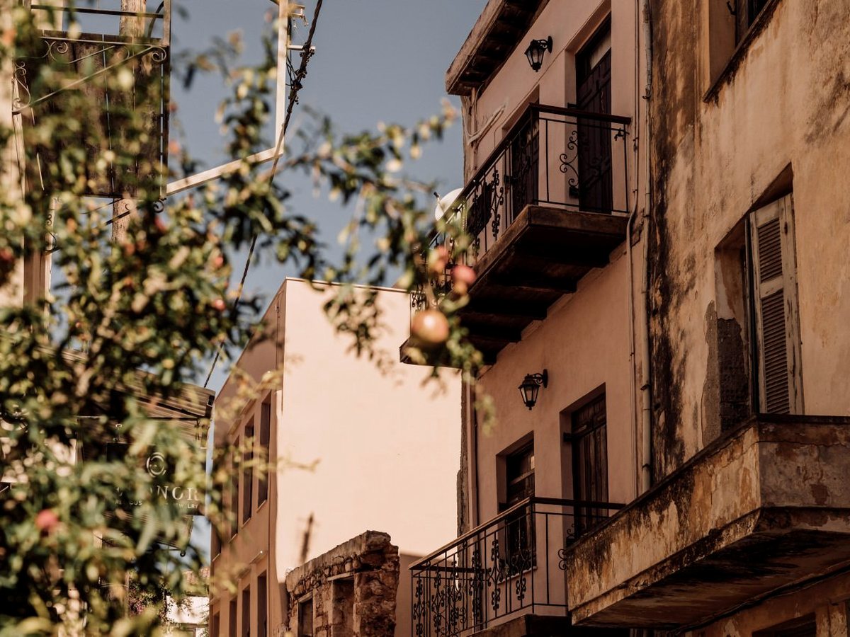 A narrow Chania old-town street of weathered Venetian and Ottoman houses with balconies and a pomegranate tree.