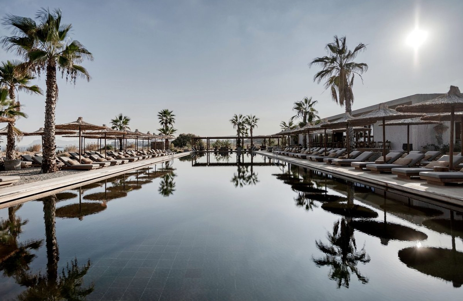 The main pool at Domes Zeen Chania, a long black-tiled pool lined with loungers and palms, with the sea beyond.