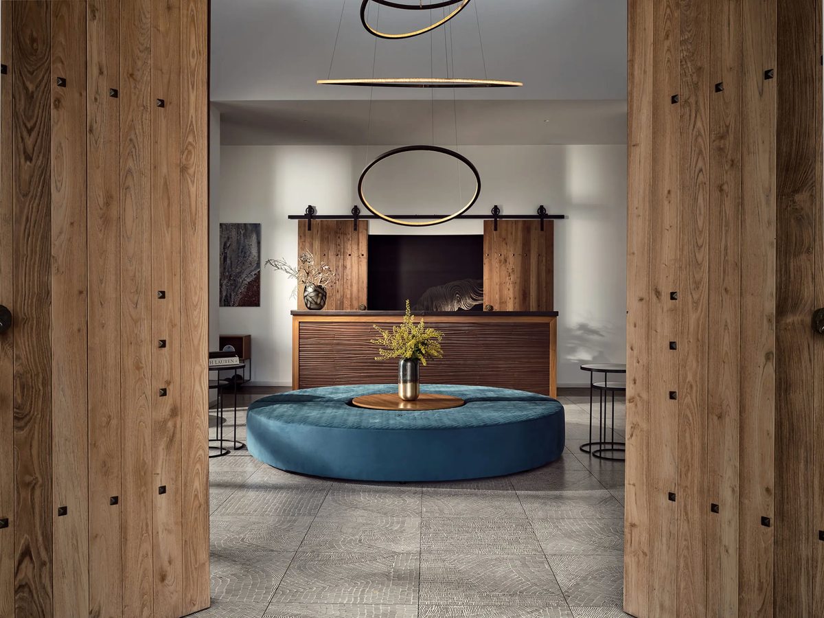 Hotel lobby with timber-panelled walls, a round blue upholstered ottoman set around a central table with a flower vase, and ring pendant lights over a grey tiled floor.