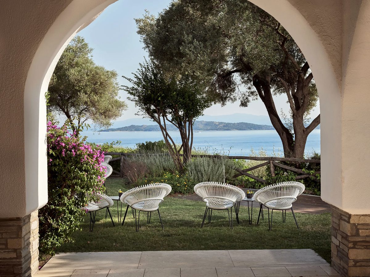Arched stone veranda with white outdoor chairs on a shaded terrace, opening onto a lawn with flowering bougainvillea and the sea beyond.