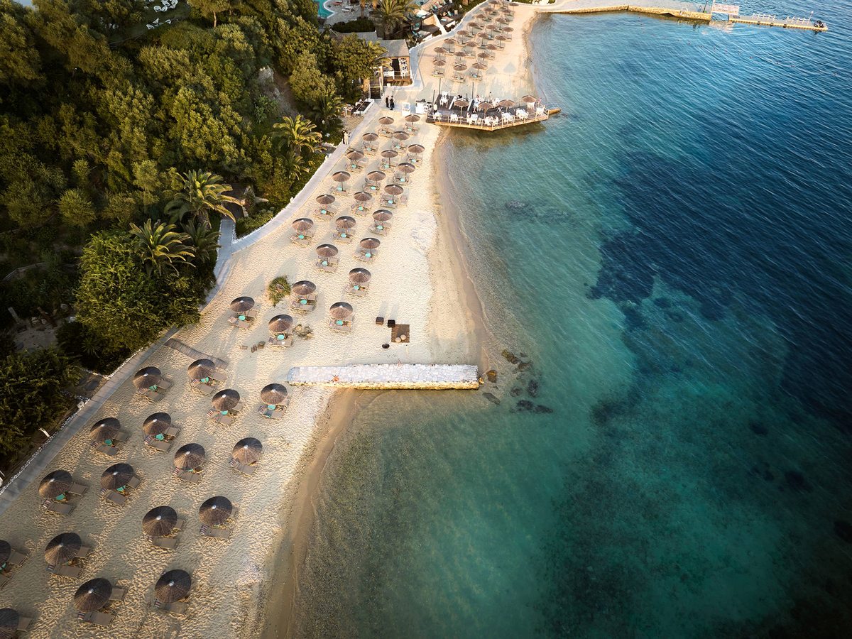 Aerial view of the private sandy beach lined with rows of straw parasols and loungers, a jetty reaching into clear turquoise water.