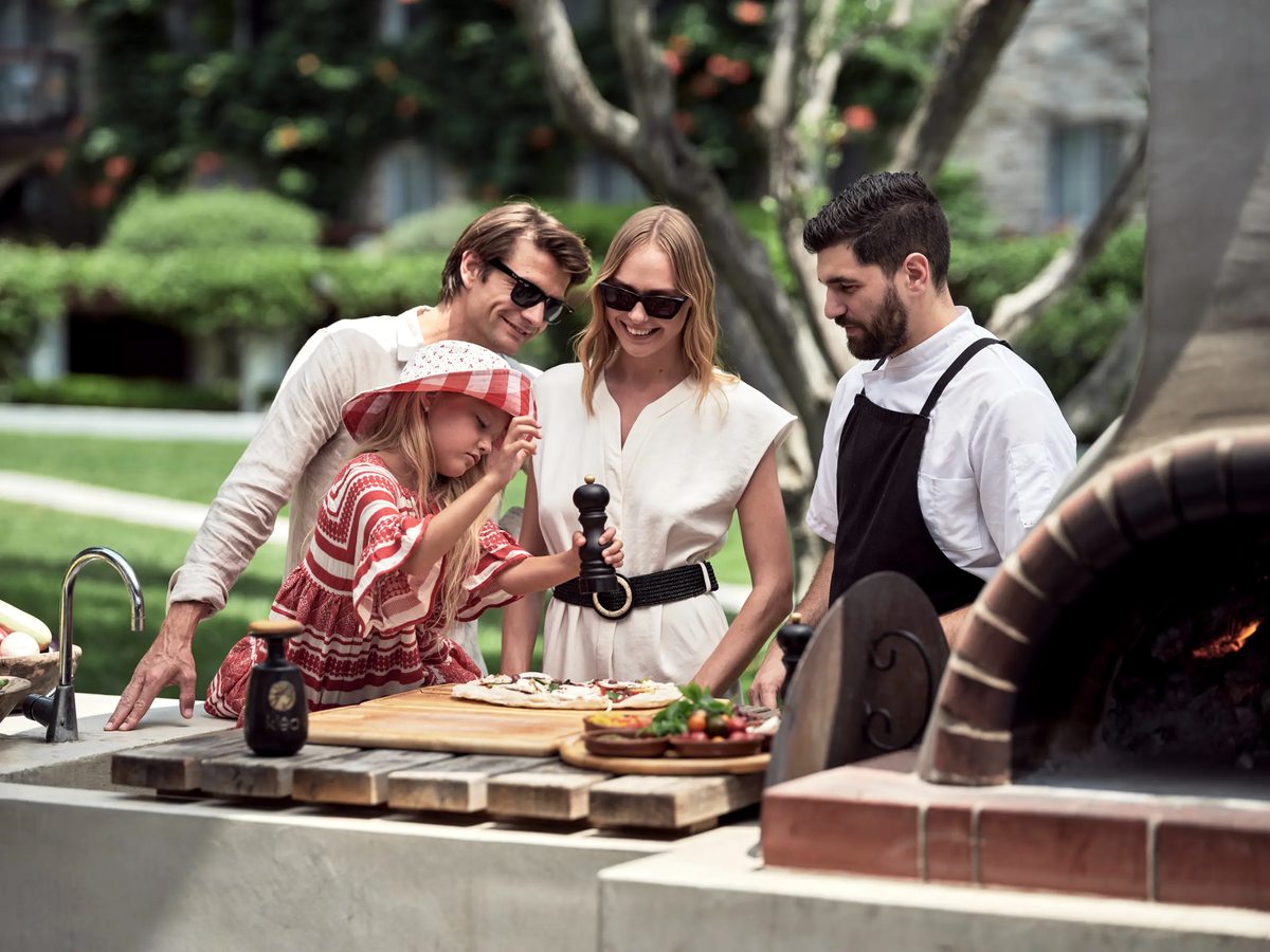 A chef at a stone wood-fired oven preparing food on a counter as a family looks on in the garden.
