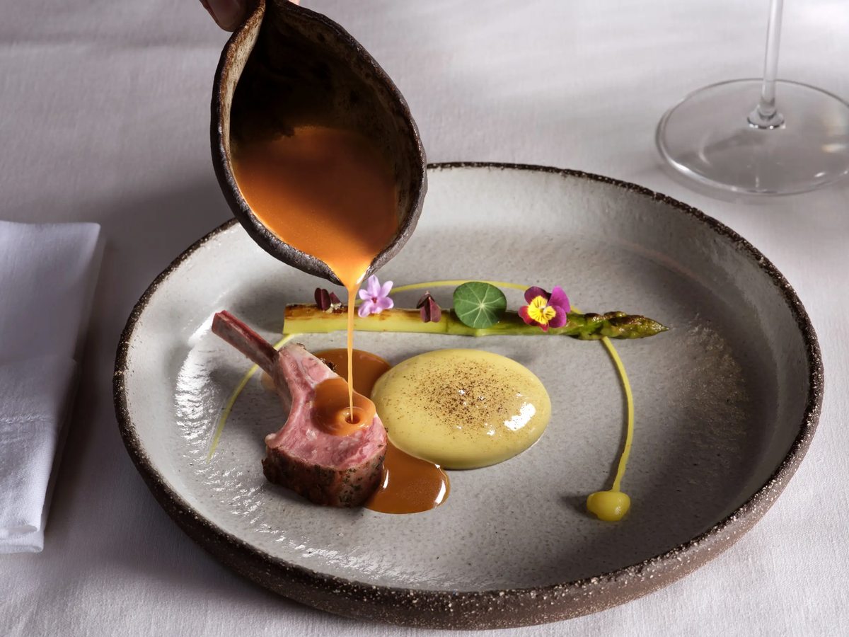 Fine-dining plate with a lamb cutlet, puree, an asparagus spear and edible flowers, with sauce being poured from a small jug.