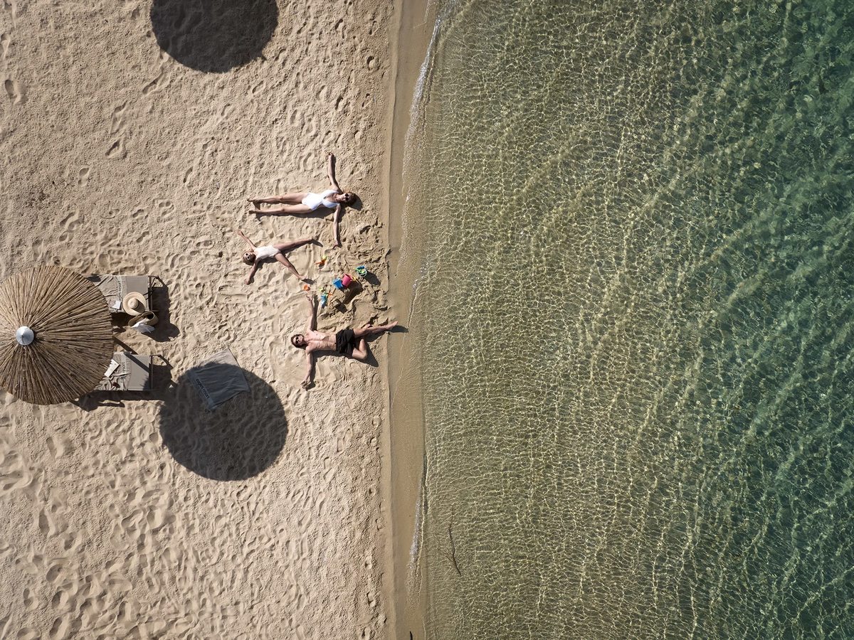 Overhead view of a family lying on the sand at the water's edge beside a straw parasol, where clear shallow water meets the beach.