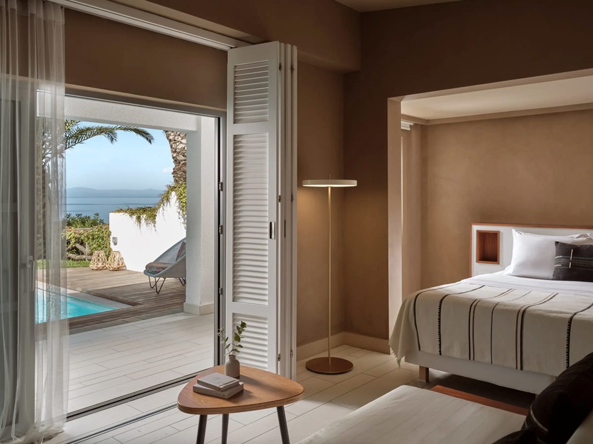 Earthy-toned suite bedroom with folding louvred doors sliding open to a terrace with a private plunge pool, a palm and a sea view.