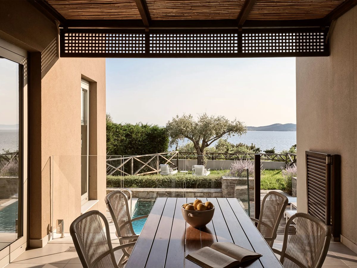 Covered suite terrace under a timber pergola with an outdoor dining table and chairs, looking past a private pool and an olive tree to the sea.