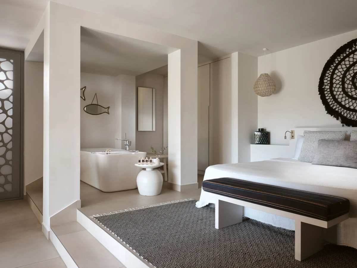 White-toned suite bedroom with a double bed and a woven wall disc, and a freestanding bath set in an open alcove behind the bed.