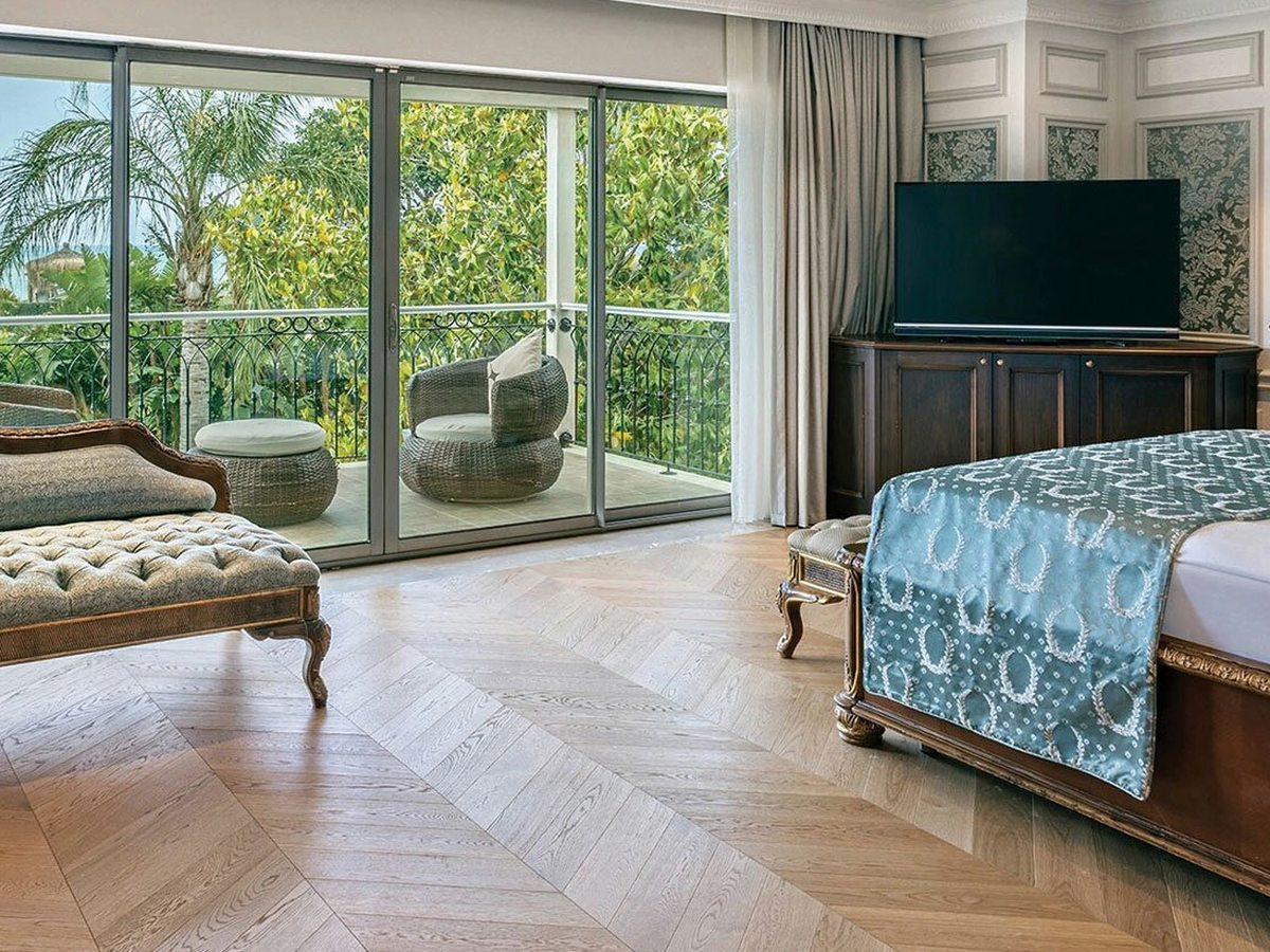A spacious suite bedroom with a large bed and chaise longue, a herringbone wood floor and sliding glass doors to a balcony with a garden view.