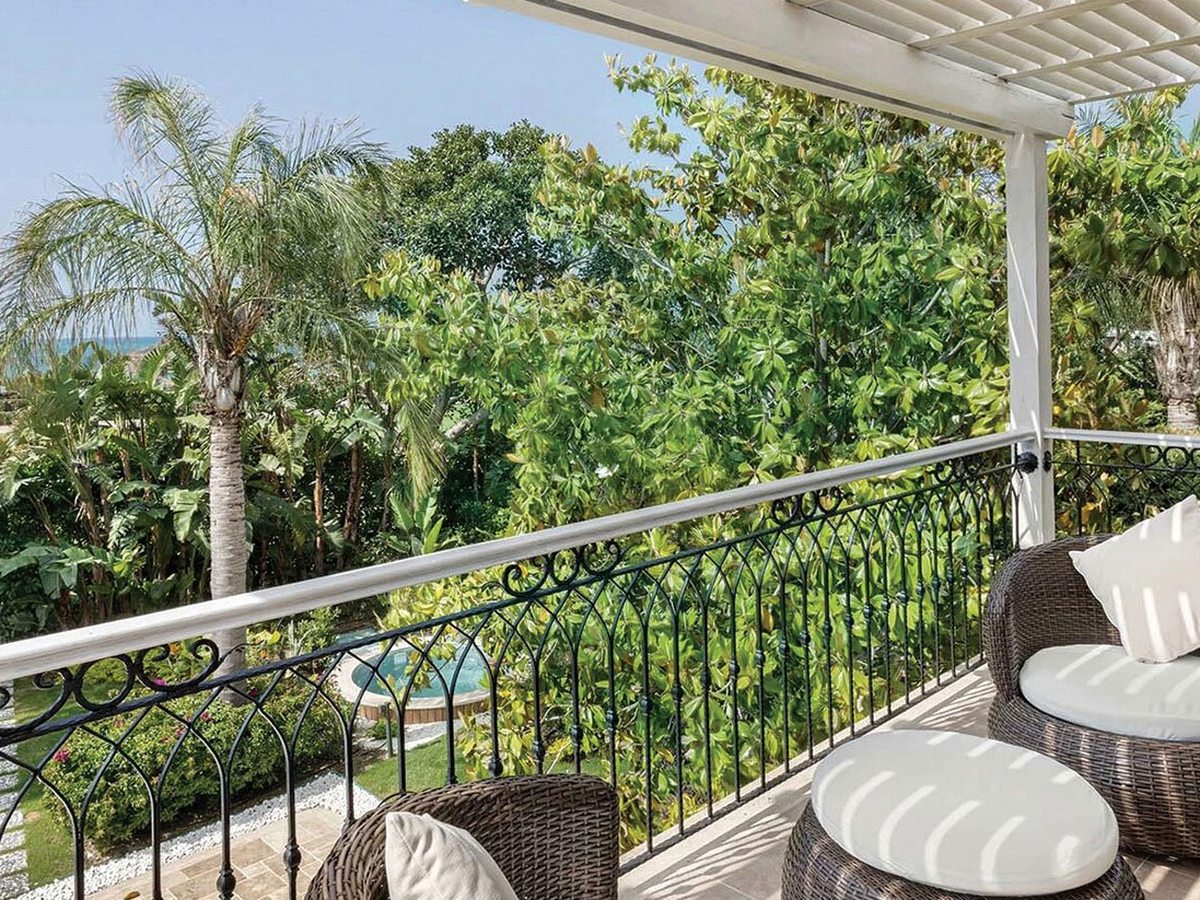 A covered balcony with wicker chairs and footstools looking out over palm-filled resort gardens toward a pool.