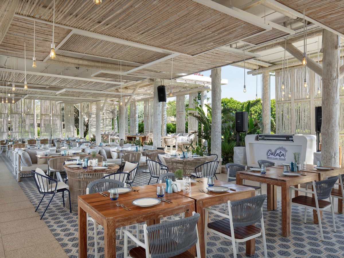 An airy whitewashed beach restaurant with a bamboo ceiling, hanging bulbs, woven chairs and wooden tables on patterned tiles, greenery beyond.