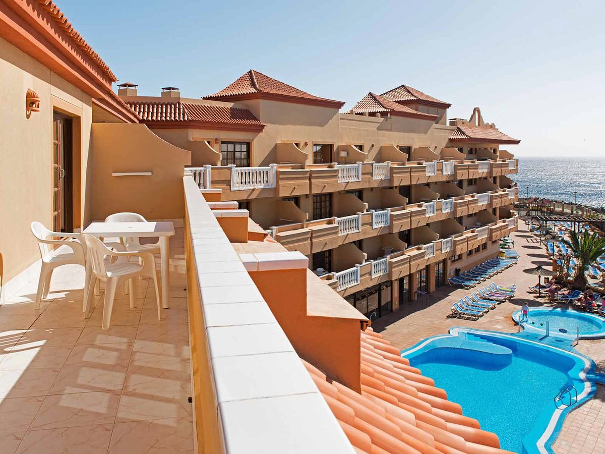 A private room balcony with a white table and chairs, looking down over the resort's swimming pool, terracotta roofs and the sea beyond.