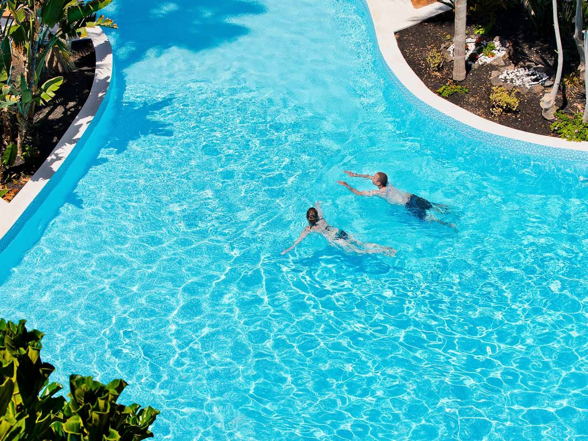 Two people swimming in a curved turquoise pool edged with green planting.
