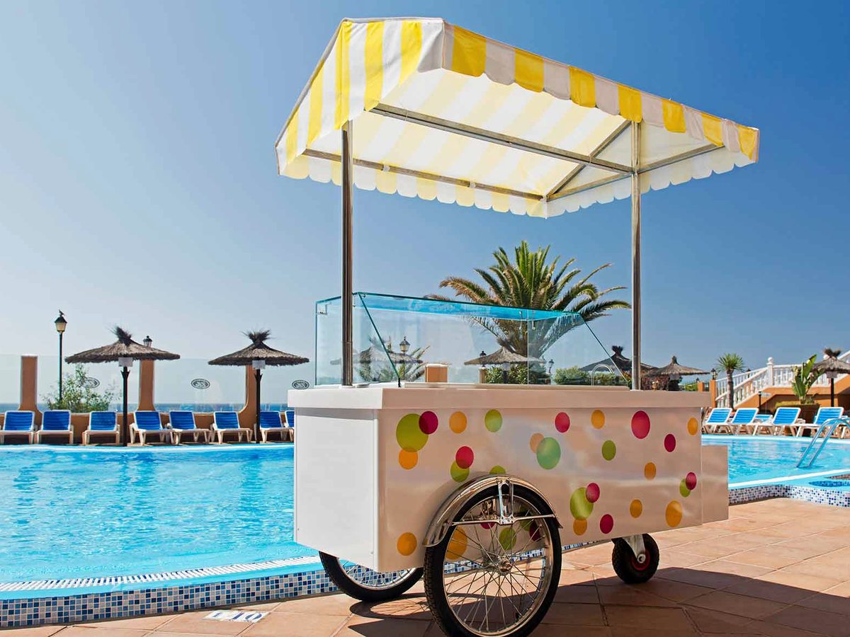 A yellow-and-white striped ice cream cart on the pool terrace, with sun loungers, parasols and palm trees behind.