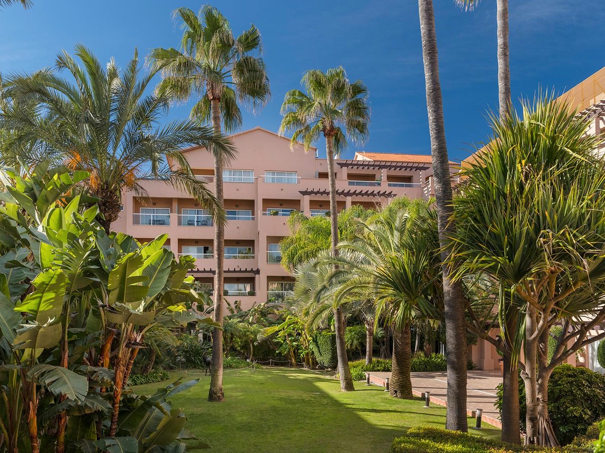 Terracotta-coloured hotel accommodation blocks with balconies set behind subtropical gardens and palms under a bright sky.