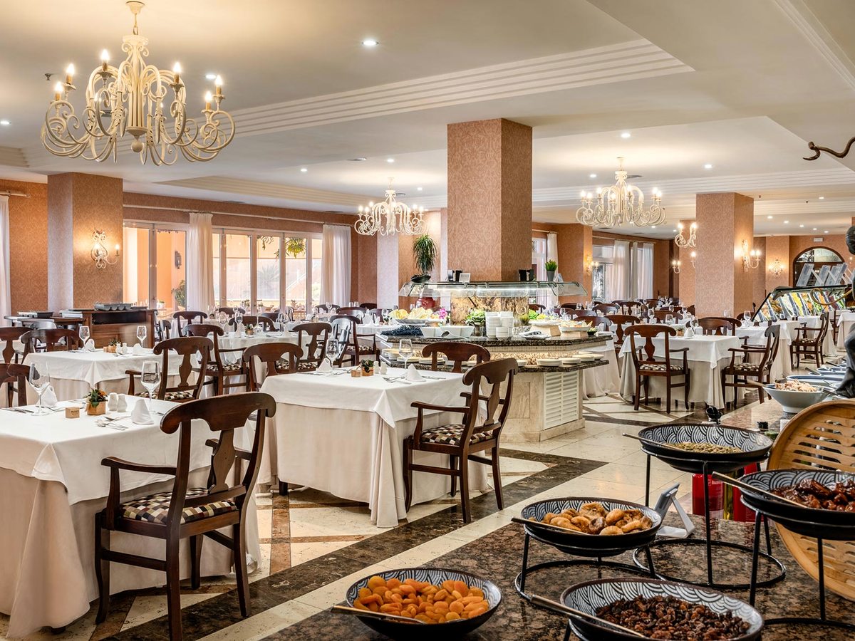 A buffet restaurant with central serving islands of hot dishes, laid tables with wooden chairs and chandeliers overhead.