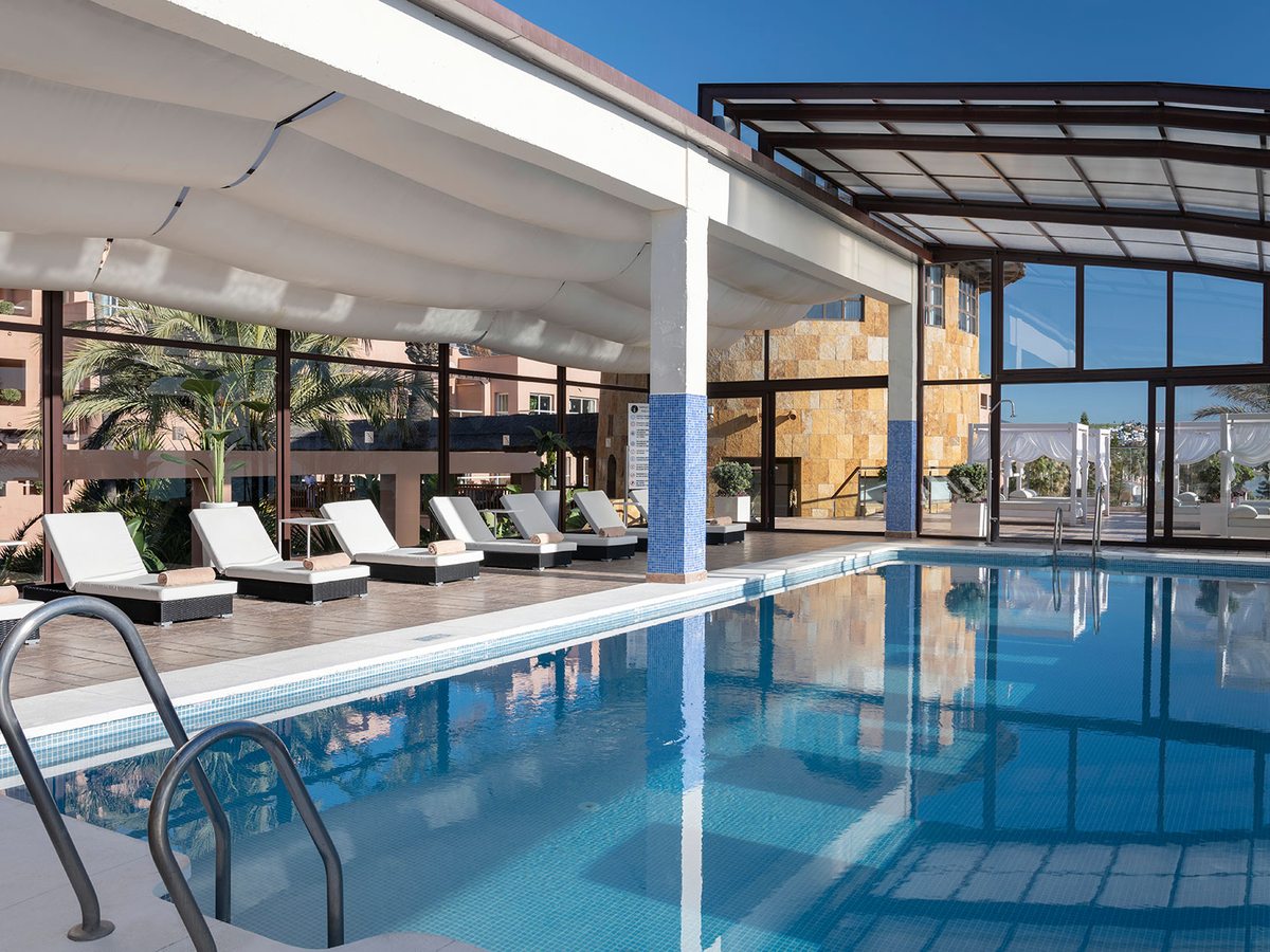 An indoor pool under a glass roof with blue-tiled columns and white sun loungers with rolled towels along the tiled deck.