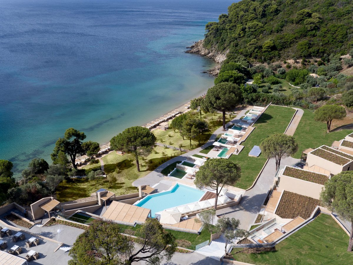 Aerial view of the resort's low sand-coloured buildings and private pools on a green slope running down through pine and olive to a sandy cove and clear turquoise sea.