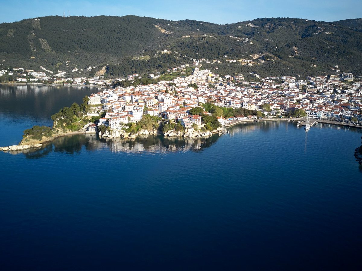 Aerial view of Skiathos Town's white houses spread around a harbour, with a wooded islet in the bay and green hills behind.