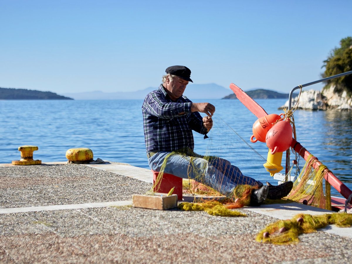 A fisherman in a flat cap sitting on a stone quay tending orange and green nets, with calm sea and a wooded island behind.