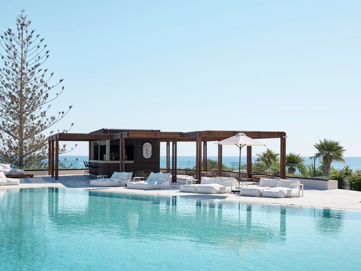 A long pool on a raised deck with white sofa-loungers, a pergola bar and the sea beyond