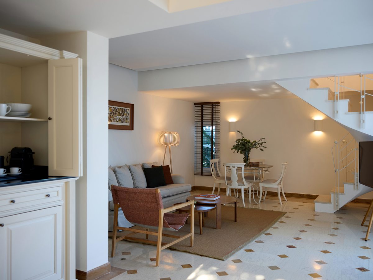 Living and dining area with a sofa, armchairs and a small dining table, a kitchenette to one side and a white staircase curving up to the floor above.