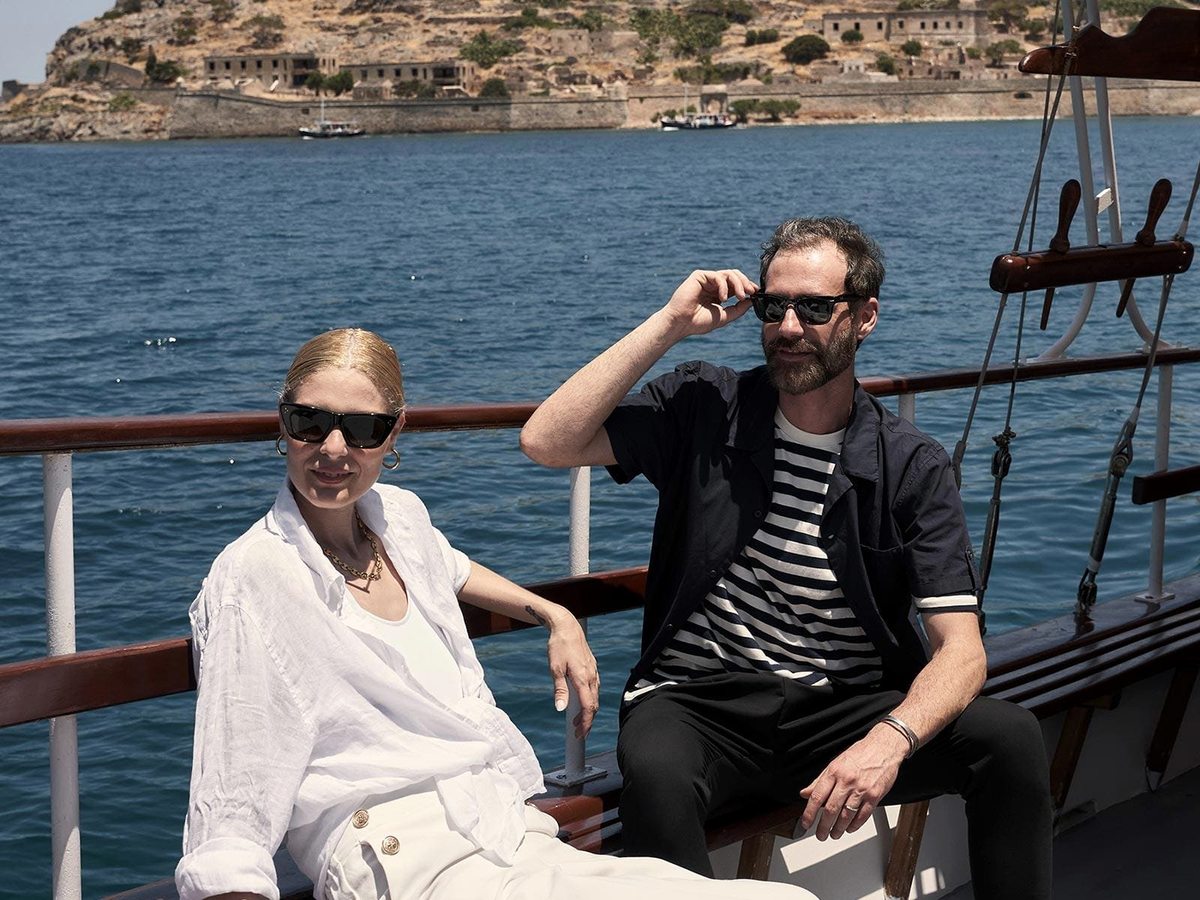 Two people in summer clothes on the wooden deck of a boat, with the fortified shore of Spinalonga behind on the blue sea.