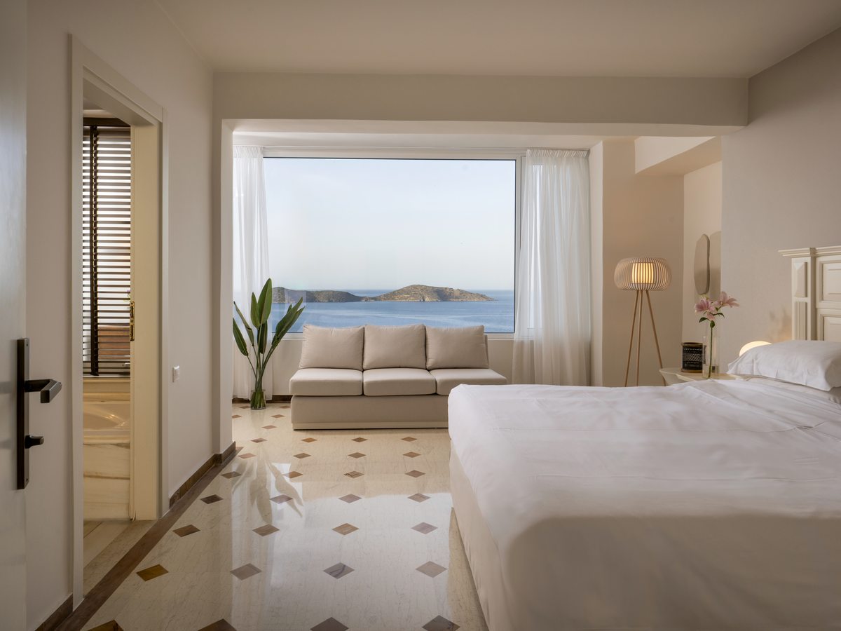Bedroom with a large picture window framing the sea and an island, a double bed, a two-seat sofa and an en-suite tub through an open door.
