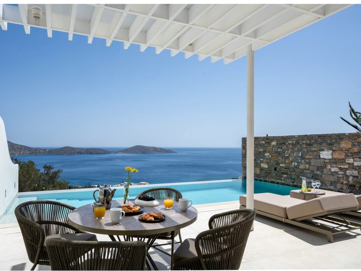Covered villa terrace with a laid breakfast table and rope chairs, an infinity pool beyond and the sea and an island on the horizon.