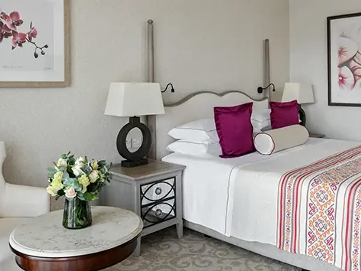 A Deluxe Room with a bed dressed in white and magenta, a mirrored bedside table, table lamps and framed orchid prints.