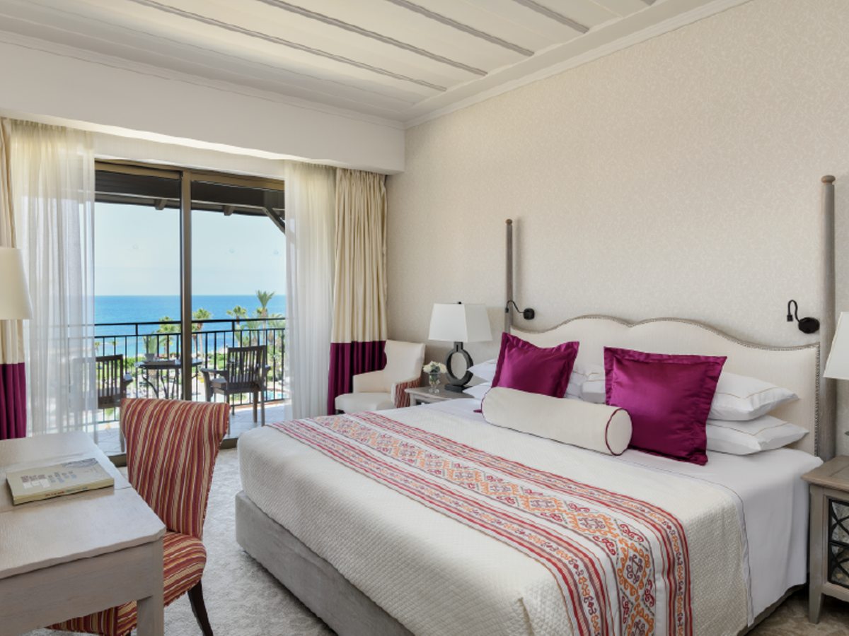 A Deluxe Room with a bed and a balcony framing a view over palm-filled gardens to the sea.