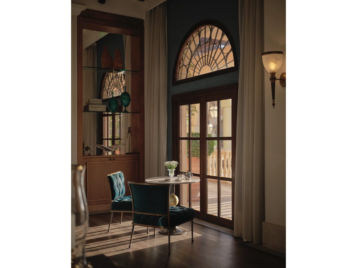 A small round marble table set for tea with two teal velvet chairs, beside a tall arched window and glazed doors opening to a sunlit terrace.