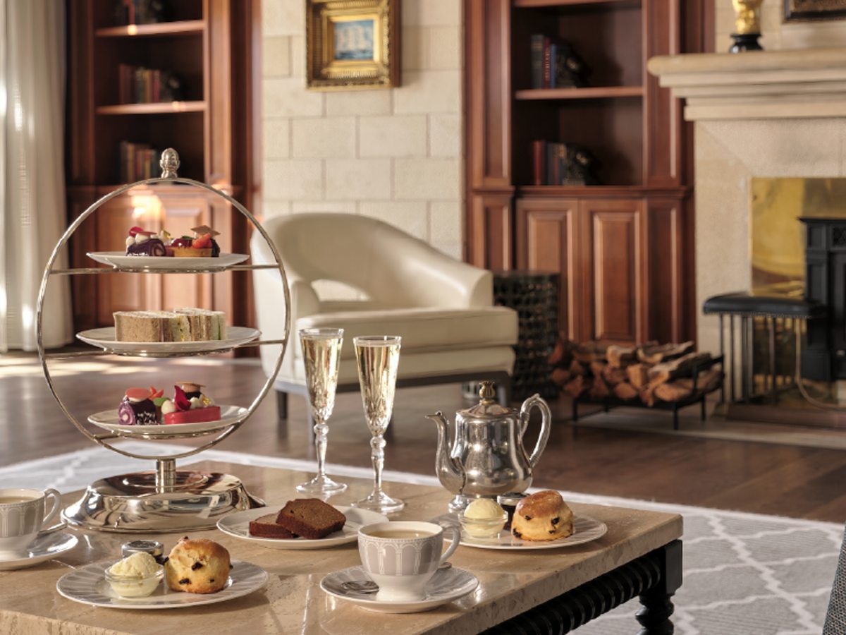 A silver three-tier stand of sandwiches, scones and pastries with tea and two glasses of sparkling wine, in a book-lined lounge with a fireplace.