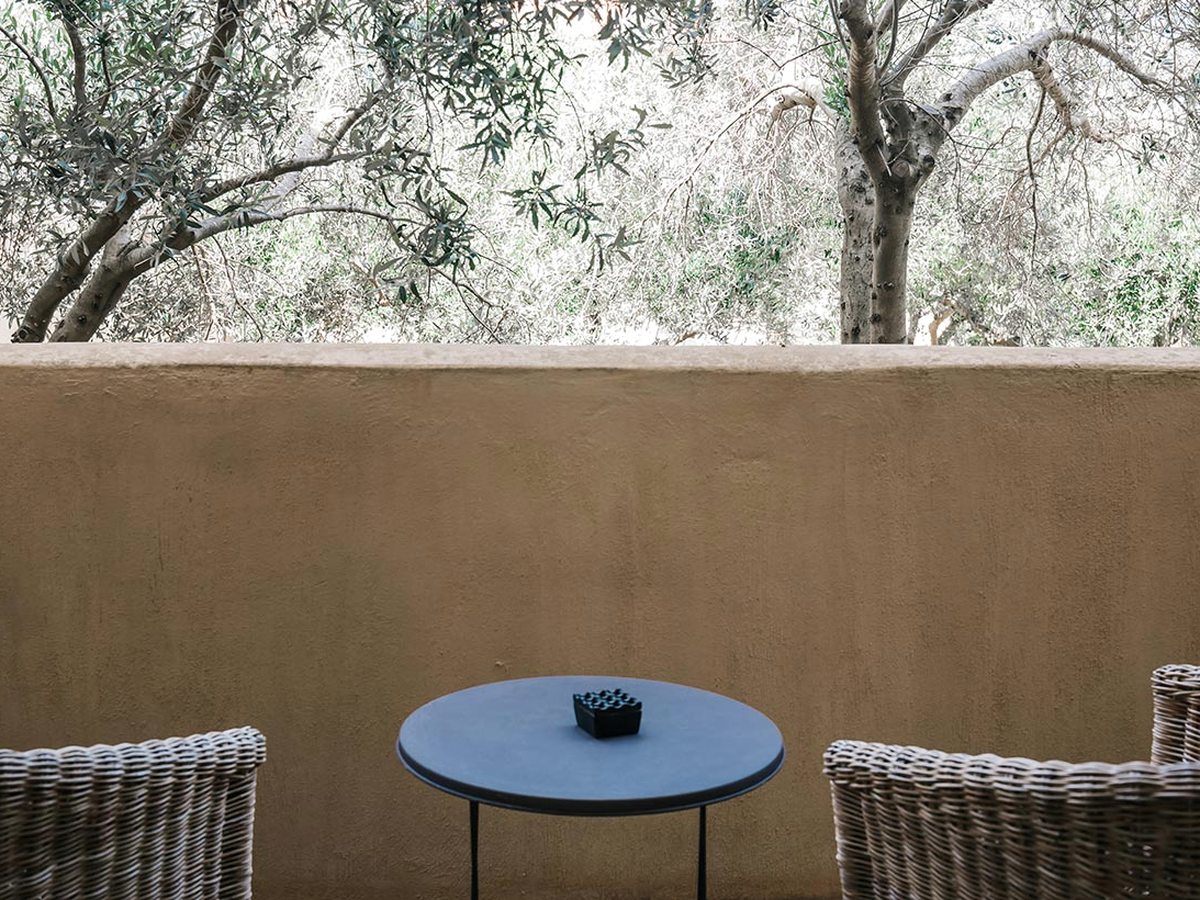 A room terrace with wicker armchairs and a small table, olive trees and a garden beyond a low wall.