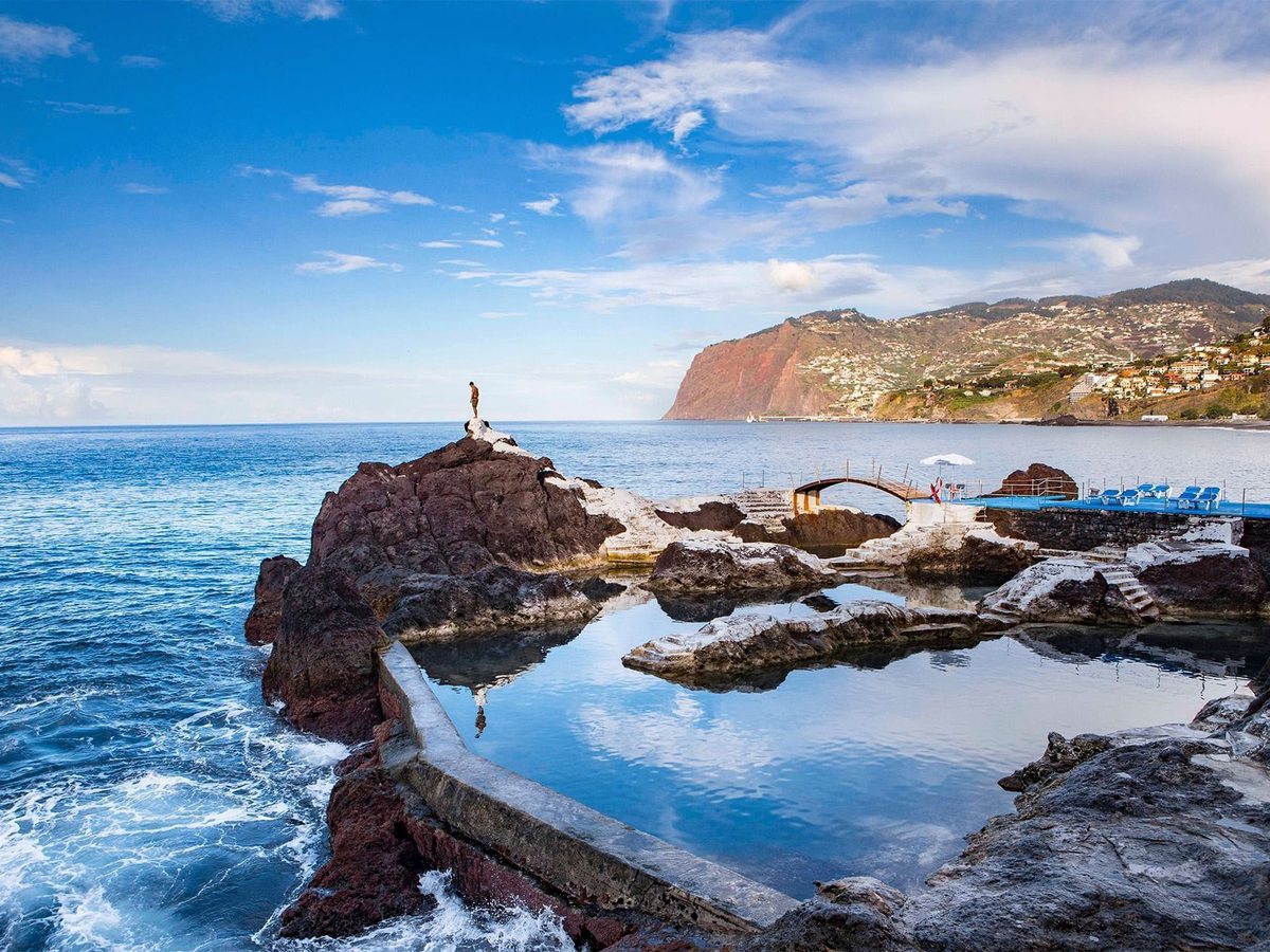 Natural rock pools formed in dark volcanic rock at the sea's edge, with a person standing on a rock, waves breaking beyond and the Funchal coastline in the distance under a bright blue sky.