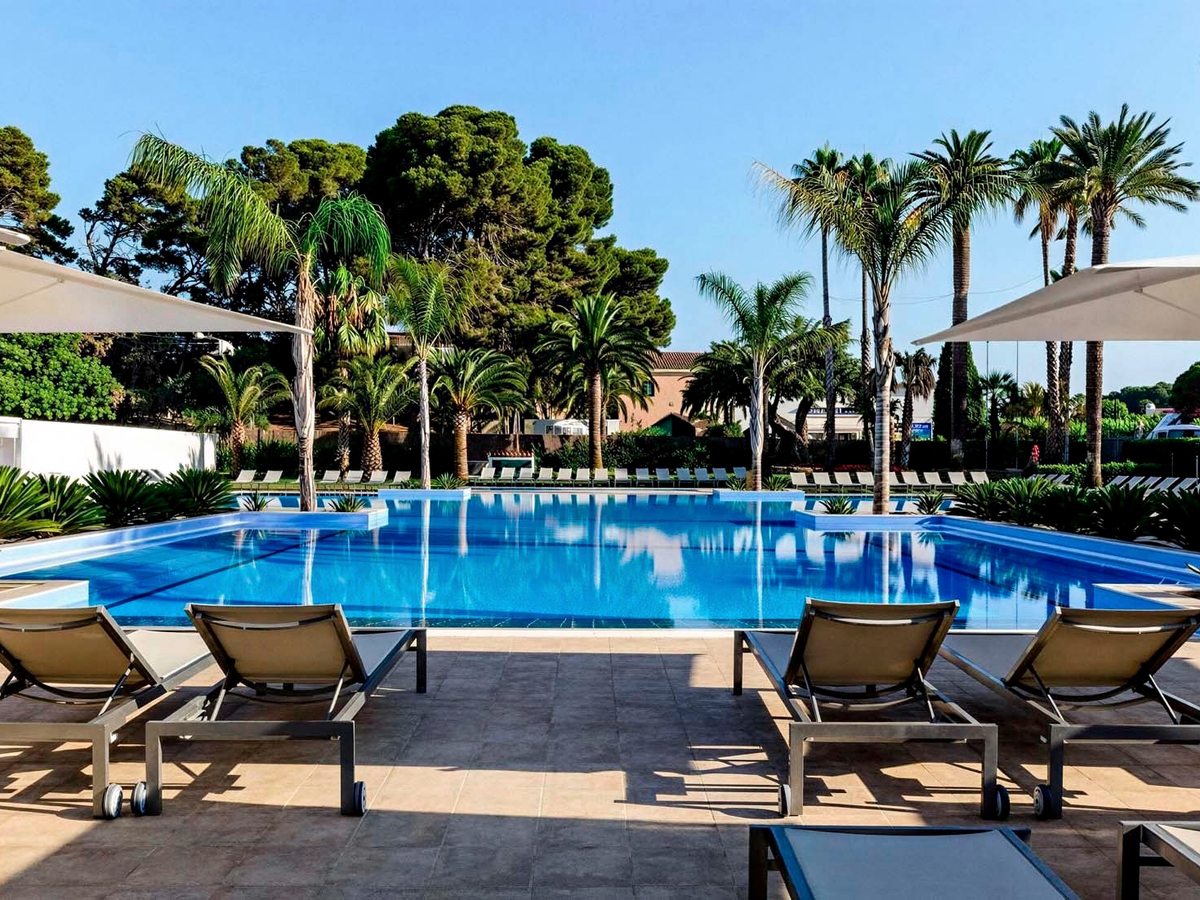 A large outdoor pool edged with sun loungers and white parasols, surrounded by tall palms and pine trees under a blue sky.