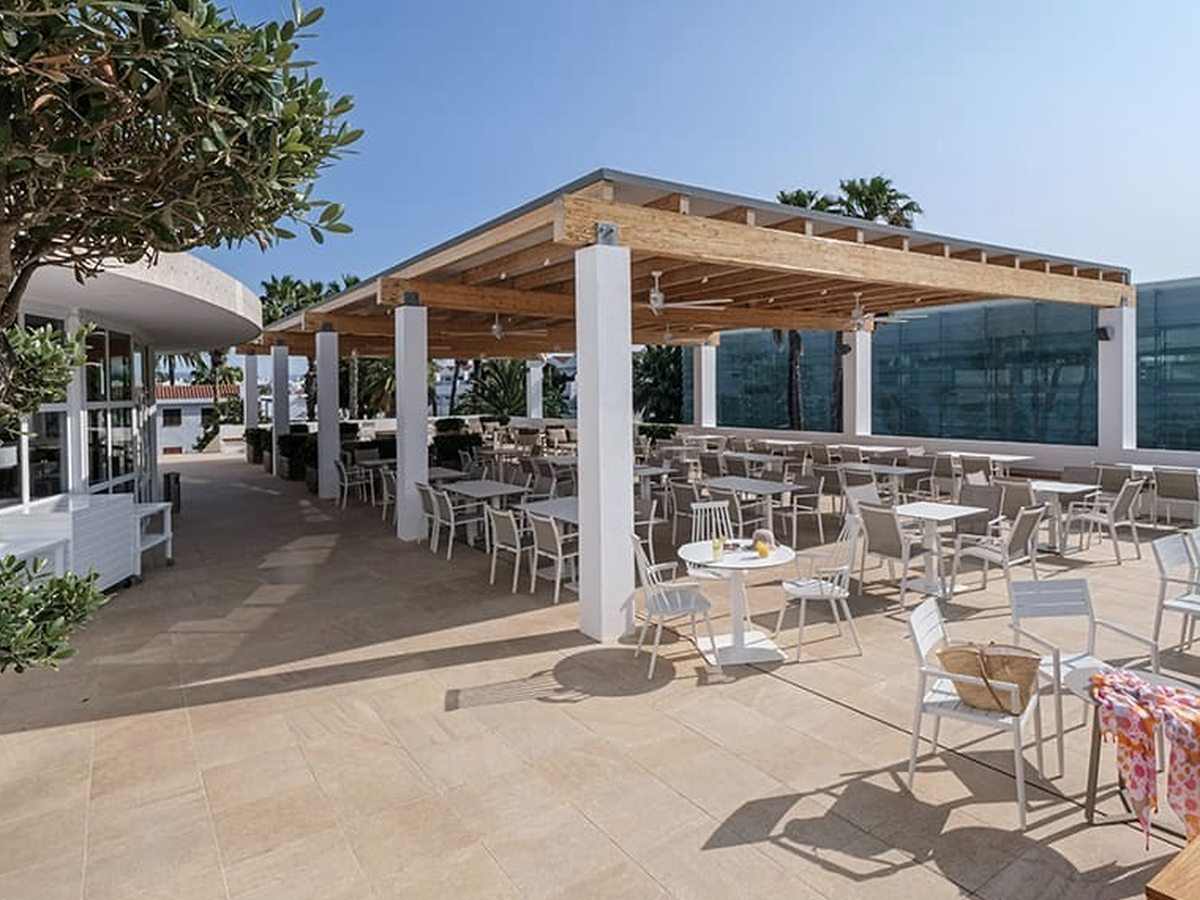 An open-air restaurant terrace with pale tables and chairs under a timber pergola, palms and low buildings beyond.