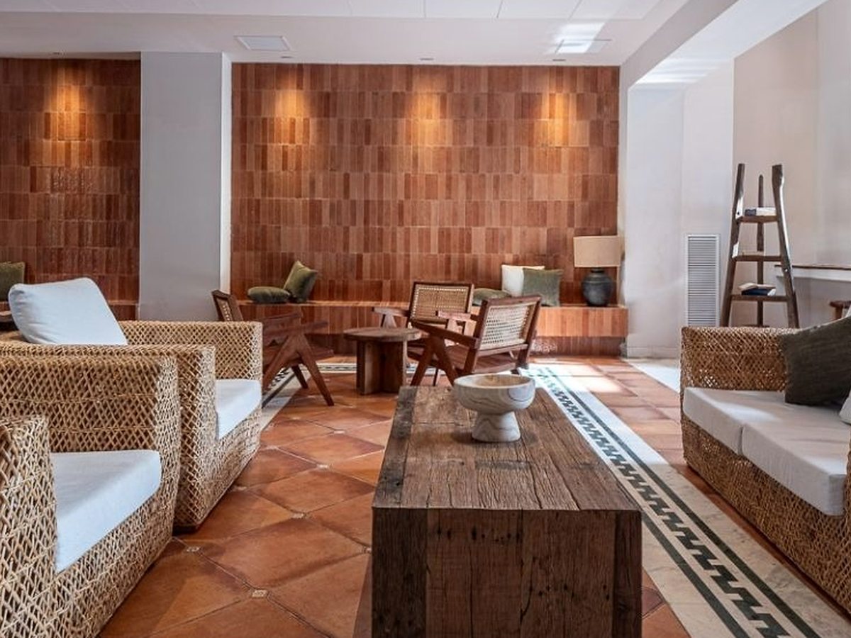 A warm lounge with woven wicker sofas, cane chairs and a rustic wooden table on a terracotta-tiled floor with a patterned border, against a wood-panelled wall.