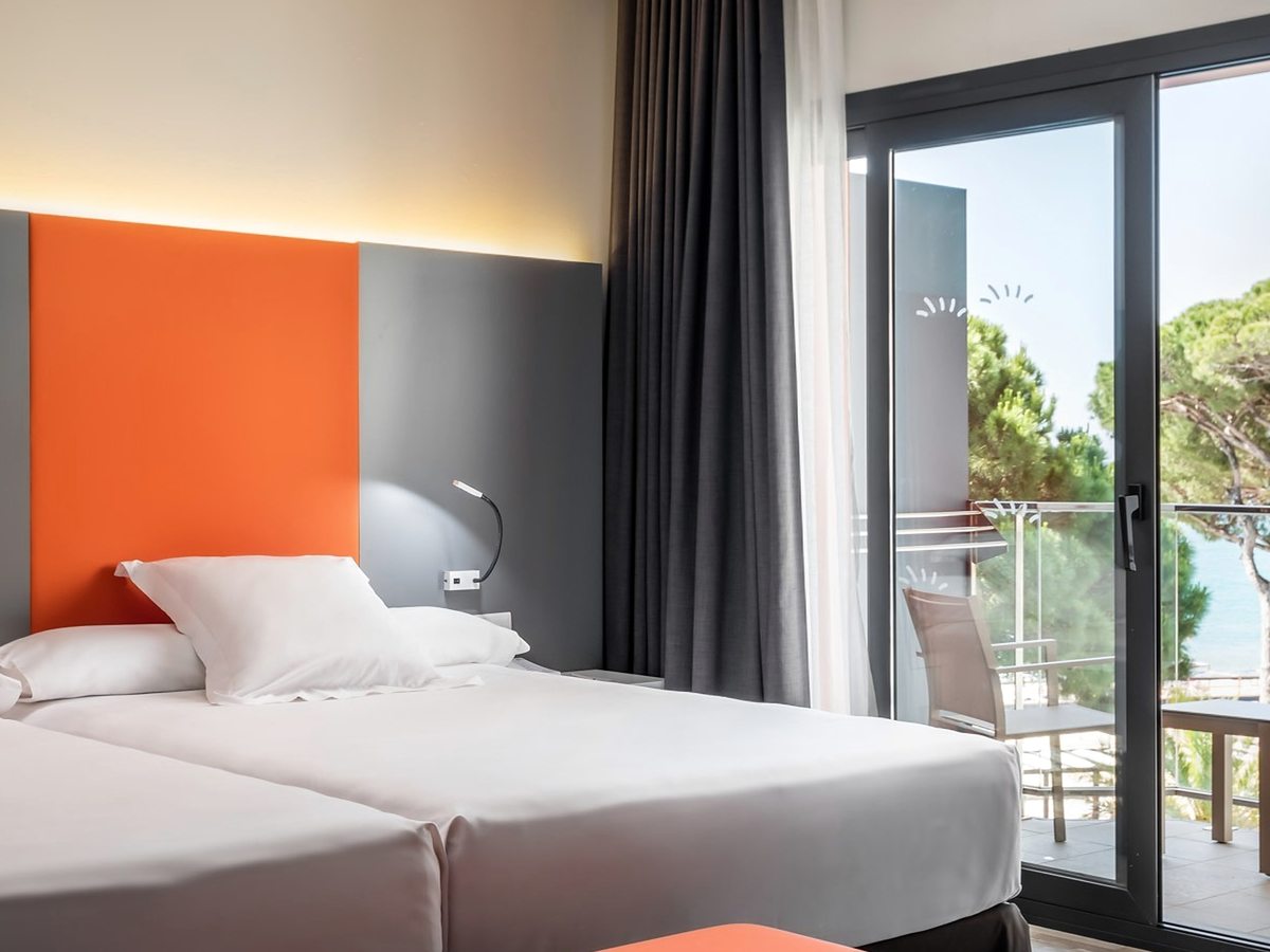 A room with twin beds pushed together, an orange and grey headboard and a sliding door to a balcony framing pine trees and the sea.