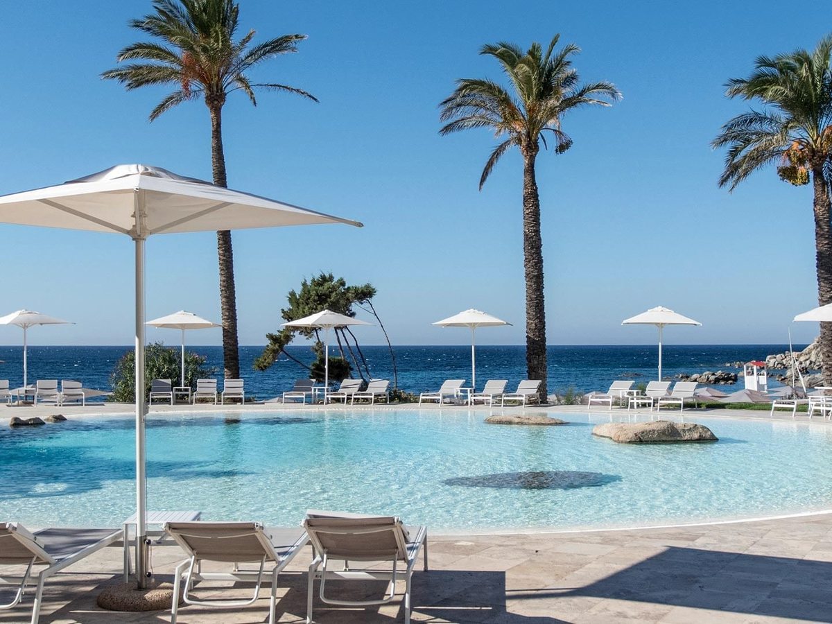 A curved outdoor pool with a central shallow area, ringed by white parasols, loungers and tall palms, the deep-blue sea behind.