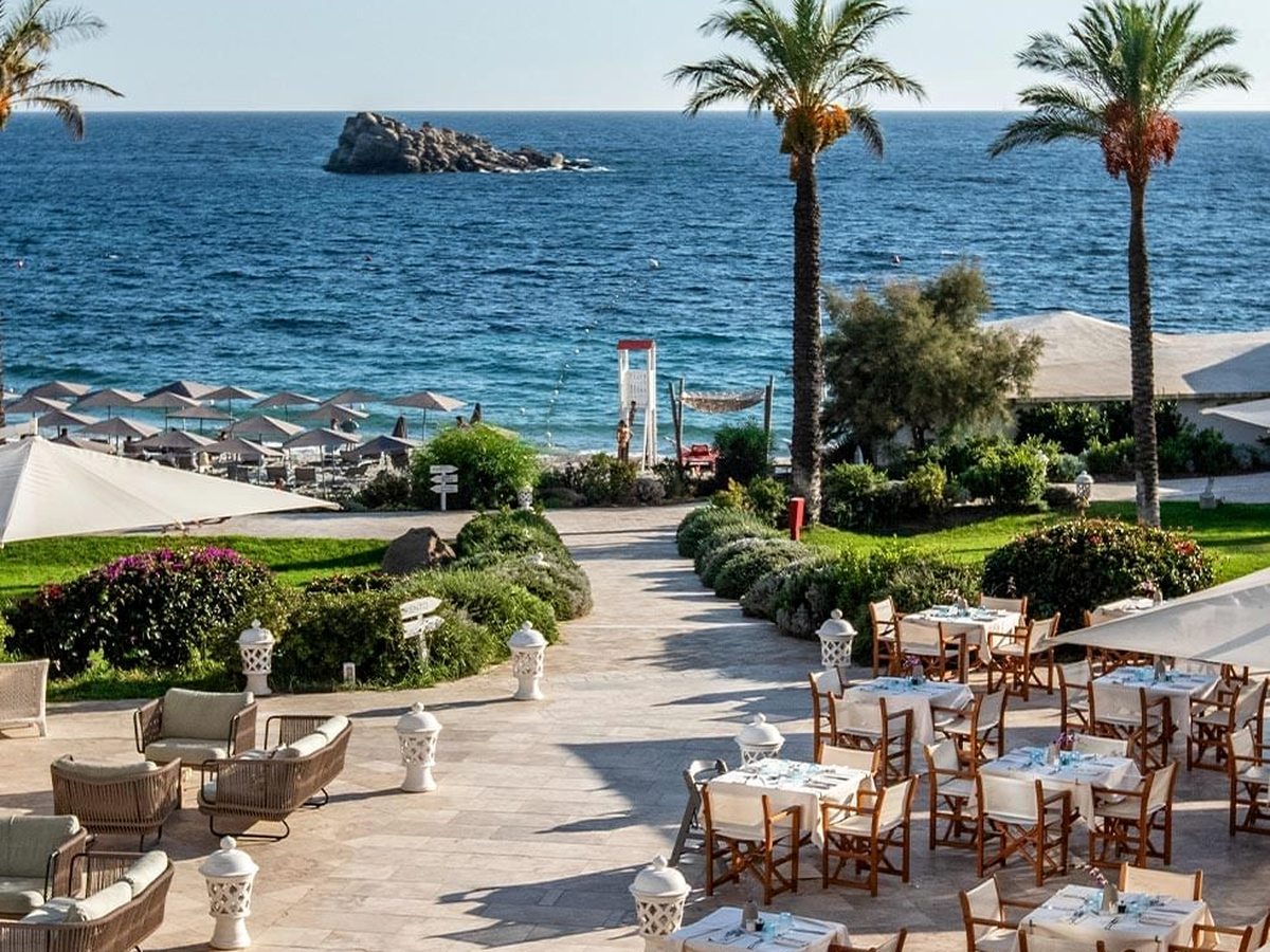Cane armchairs and set tables on a paved terrace among flowering shrubs and palms, looking down to beach umbrellas and a rock stack offshore.