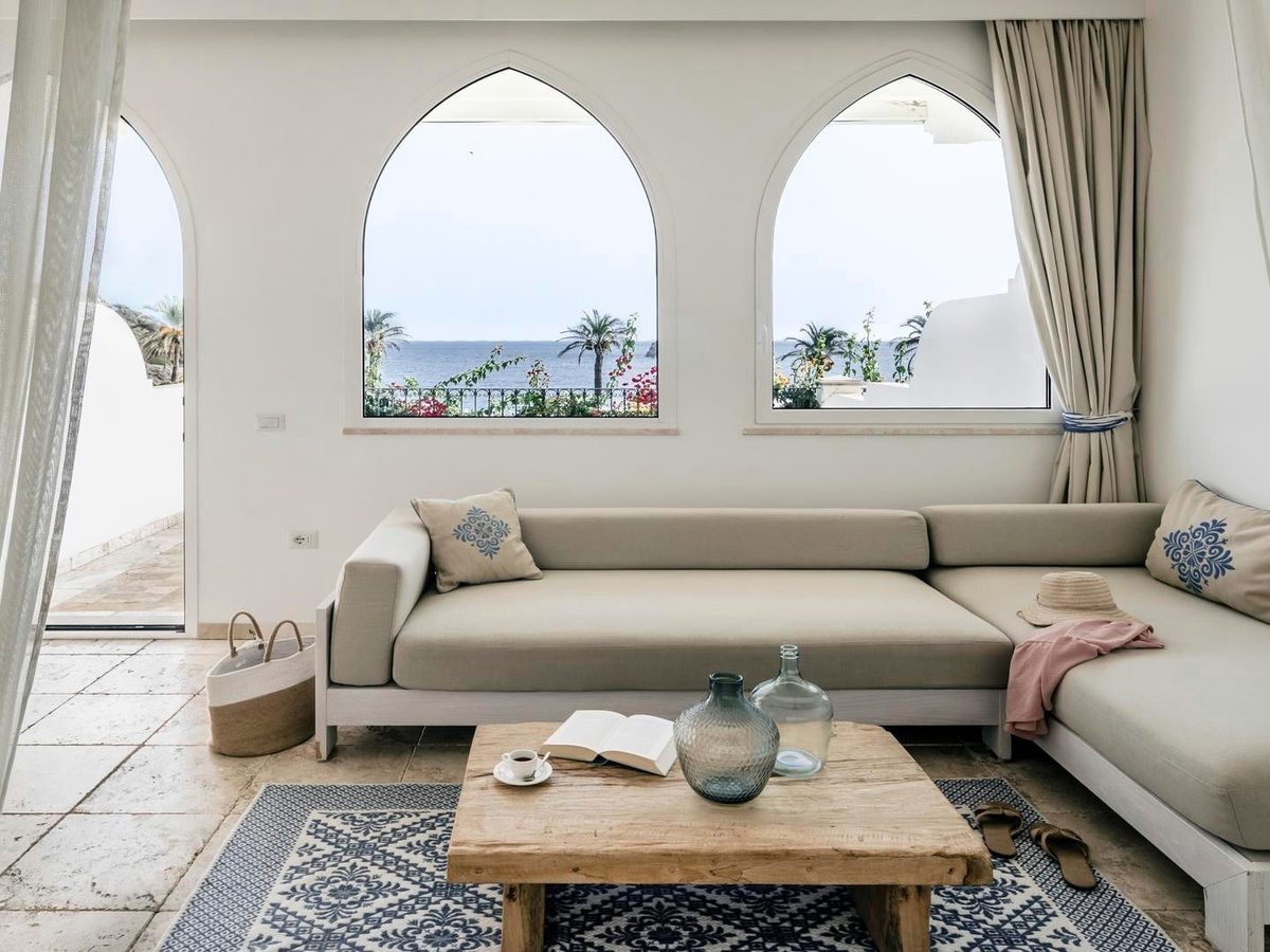 A long pale sofa below three arched windows framing the sea, with a low wooden coffee table and a patterned rug.