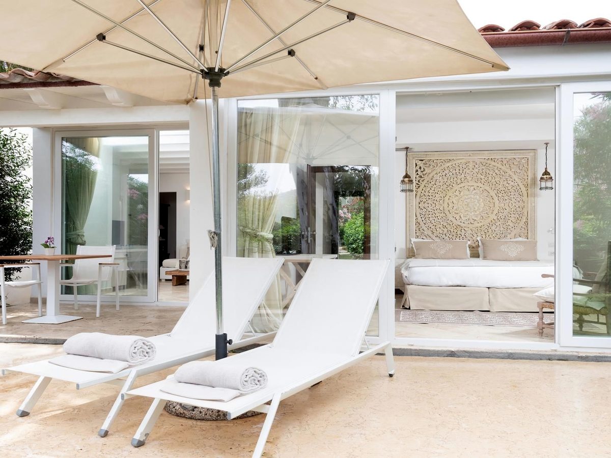 Two white sun loungers with rolled towels under a parasol on a travertine terrace, glass sliding doors behind opening into a bedroom.