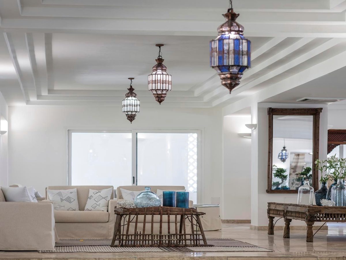 A long lobby with pale linen sofas, hanging Moorish lanterns and an arched doorway to a bright terrace at the far end.