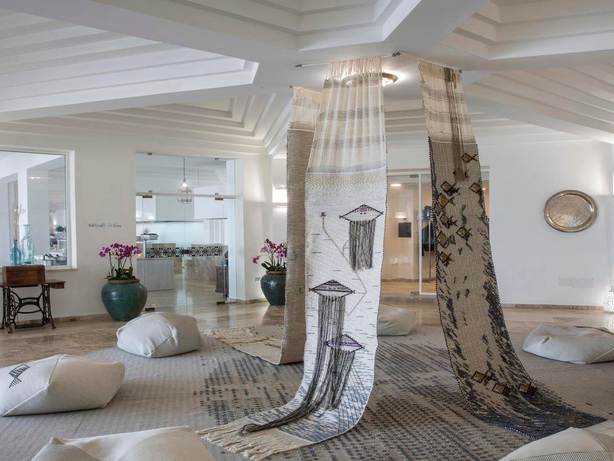 A lounge with white floor cushions on a patterned rug, tall woven textile hangings draped between two columns and a dark carved wooden console.