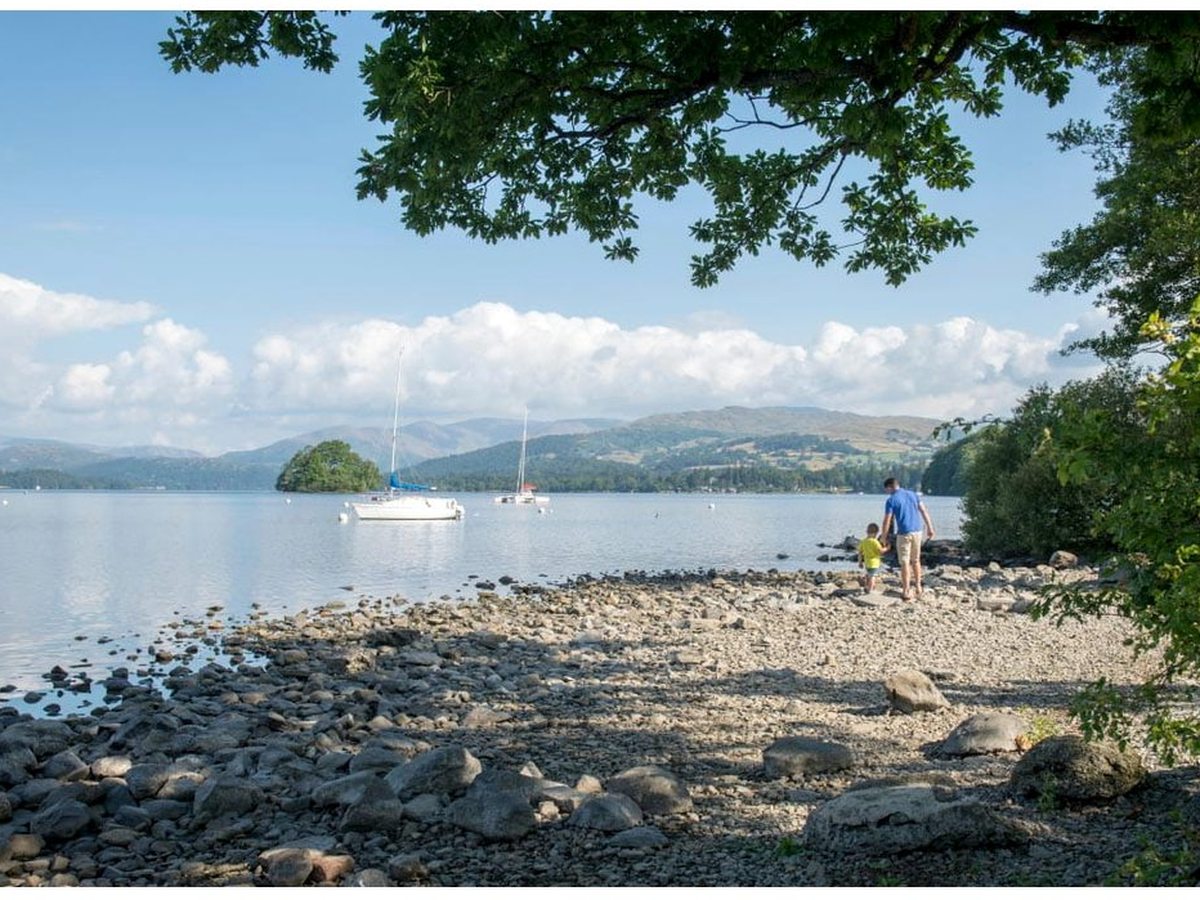 A rocky lakeshore beneath overhanging trees, sailing boats moored on calm water and the Lakeland fells in the distance, with a parent and young child walking the stones.