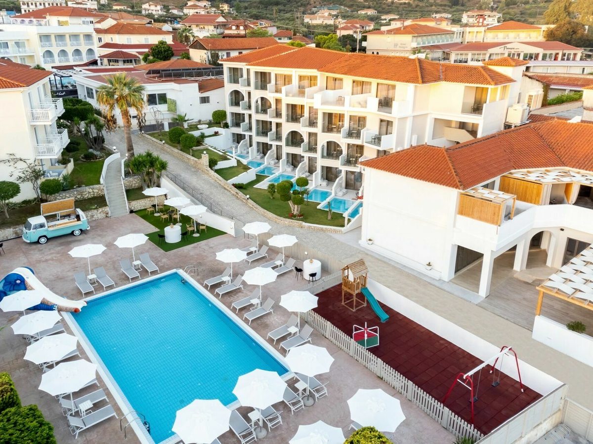 Aerial view of a rectangular pool with white parasols and loungers, a small children's climbing frame and slide on a red safety surface beside it, and a room block behind.